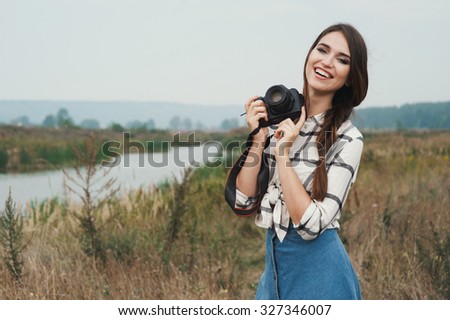 Cute countryside lady with brown hair posing against ranch house and pond with camera. She stands near tall grass and reed against rural scape. She wears jeans dress. Camera is black