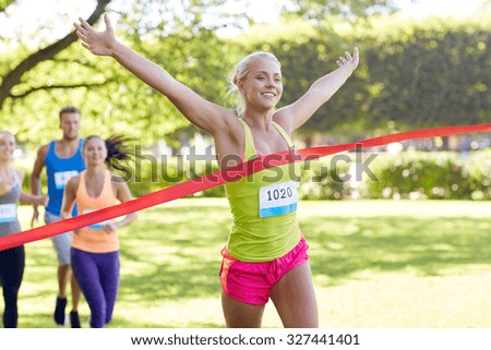 fitness, sport, victory, success and healthy lifestyle concept - happy woman winning race and coming first to finish red ribbon over group of sportsmen running marathon with badge numbers outdoors Royalty-Free Stock Photo #327441401