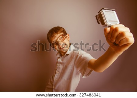 teenager boy brown hair European appearance in sunglasses holding a camera action and makes self on a gray background, laughing, fooling around, shooting retro