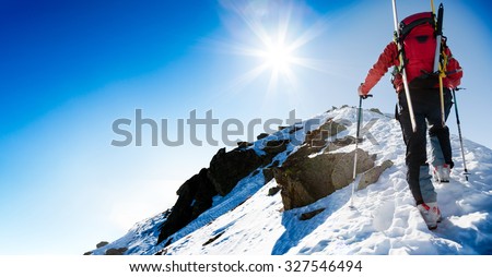 Mountaineer walking up along a snowy ridge with skis in the backpack. In background blue sky and shiny sun. Concepts: adventure, determination, extreme sport. Large copy-space on the left for text.