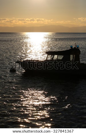 Marine sunset scene with lights reflecting at dark seaways in foreground and sun with halo glade shines over horizon in dusk against clouds background with boat silhouette, vertical picture