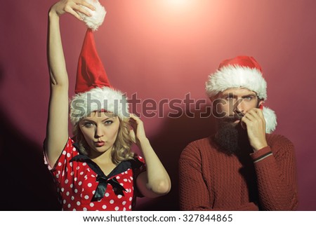 New year funny couple of blond woman with curly hair and man with long beard in red santa claus hat celebrating christmas standing on studio purple background, horizontal picture