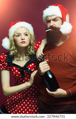 New year couple of blond woman with curly hair and man with long beard in red santa claus hat smoking cigarette holding wine bottle celebrating christmas standing on studio purple background, vertical