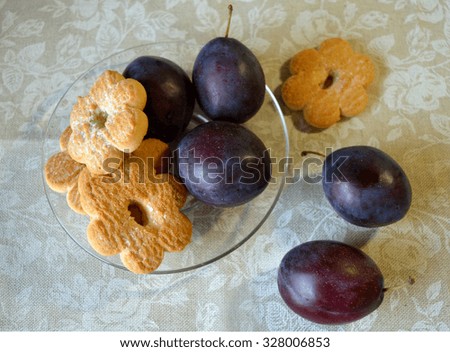 Cookies in the shape of flowers and plums on the transparent saucer and on tablecloth with white roses