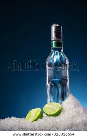 Bottle of vodka and two lime halves with water droplets on a surface in cold ice on blue gradient background. Focus on a bottle.