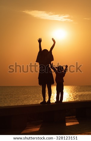 Mom and little daughter with their hands up to meet the sunrise over the sea. Silhouettes of women and girl. Toned image.