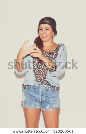 Modern redhead teenage girl with cap taking a selfie on smartphone. Happy young woman in denim and leopard print using smartphone. Studio shot, vertical, matte look, medium retouch.