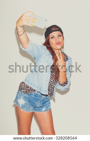 Fashionable cool redhead teenage girl in denim shorts and jacket taking a selfie gesturing two fingers. Studio shot, vertical, matte look, medium retouch.