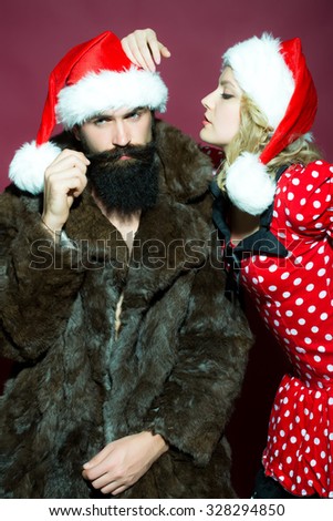 New year young couple of blond woman with curly hair in dress and man with long beard in red santa claus hat and fur coat celebrating christmas standing on studio purple background, vertical picture