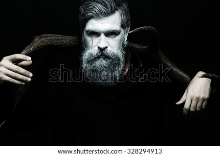 Portrait closeup view of one halloween holiday celebration character senior man with spooky painted face of zombi and long white lush beard holding animal antlers black and white, horizontal picture