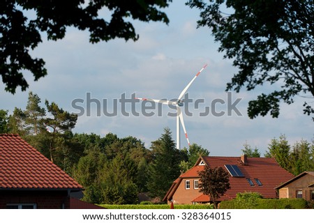 View of one big producing energy wind mill with propeller in village with houses among green lush trees on blue sky natural background, horizontal picture