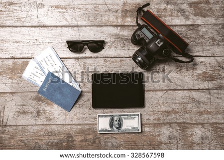 Top view photo of professional camera, tablet computer, passport, tickets, sunglasses and money. Objects are on light colored wooden floor