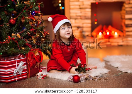 a little girl with brown hair in a New Year's cap sits near a Christmas tree Santa with gifts in red dress barefoot in a New Year's interior with a fireplace smiling and looking away