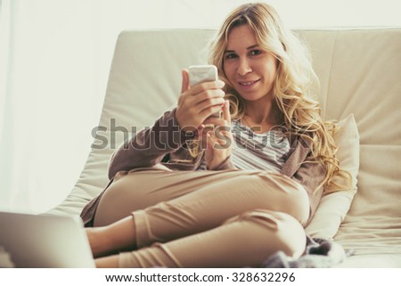 Happy young woman is relaxing on comfortable couch and using mobile phone at home. Photo toned.