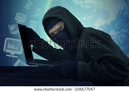 Image of male robber wearing mask and looking at the camera, stealing laptop computer and business information