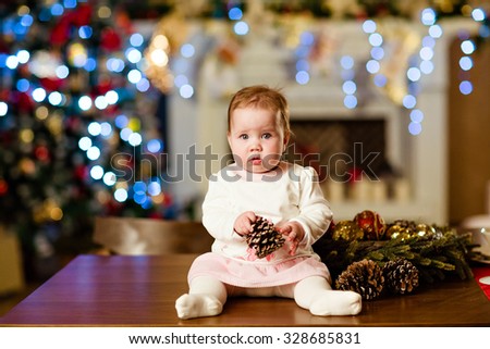 Chubby little cute baby girl in white dress sitting on the table in the New year, the Christmas lights and fireplace