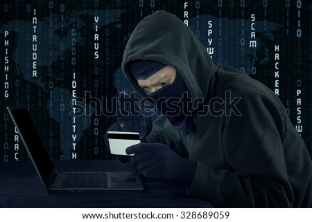 Image of male villain wearing mask and using laptop computer while holding credit card