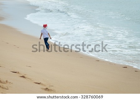 Picture of exciting little kid in Santa hat running on seashore beside waves. Backview of child in front of swash on sunny seaside background.