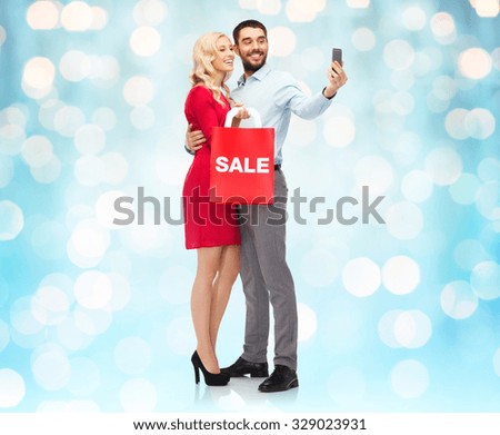 people, couple, sale, technology and holidays concept - happy young woman and man with red shopping bag taking selfie by smartphone over blue lights background