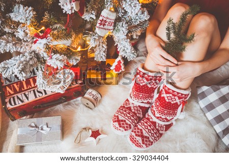 Young pretty woman in warm cozy clothes is sitting on the light wooden floor in her bright house near the christmas tree and holds a branch of fir in her hands.