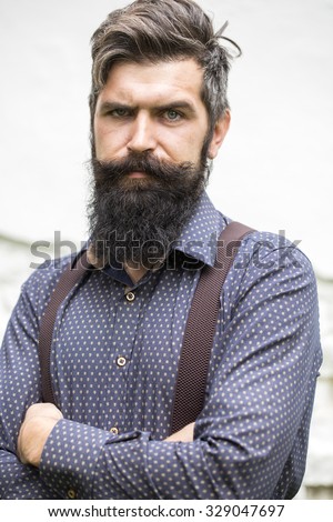 Closeup view of one handsome senior stylish man with black hair and long lush beard in blue shirt standing outdoor on white wall background, vertical picture