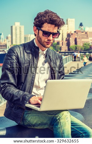 European businessman traveling, working in New York. Wearing black leather jacket, blue jeans, sunglasses, a guy with beard, sitting on bench at deck on harbor, reading, working on laptop computer. 
