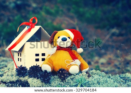 House of paper gift with red ribbon with soft Teddy bear in a Santa hat in the forest on the background of moss and cones. Concept of sale or purchase house. Greeting card for Christmas or new year