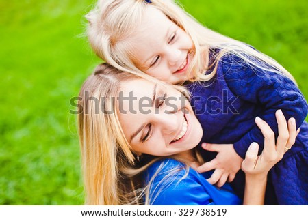 Young beautiful blonde mother close eyes and hug her little daughter smiling and relaxing in park in summer. Both wear blue dresses and laugh happily. Concept of happiness in parenthood and family