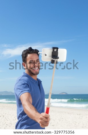 Happy guy with blue shirt and beard at beach taking selfie with stick