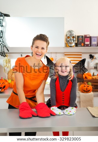 Happy young mother with daughter in bat costume wearing oven mitts standing in Halloween decorated kitchen. Ready to halloween party. Traditional autumn holiday