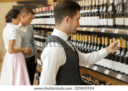 Portrait of a handsome bearded sommelier standing in a shop taking a bottle from the shelf, couple choosing a bottle of wine on the background, selective focus