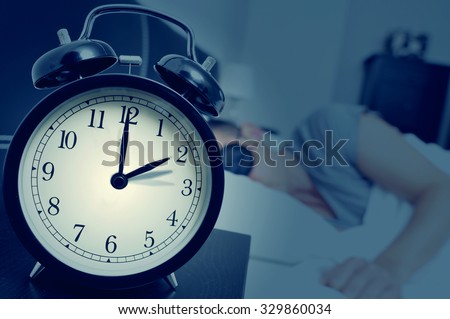 closeup of an alarm clock on a nightstand adjusting backward one hour at the end of the summer time, while a young man sleeps in bed Royalty-Free Stock Photo #329860034