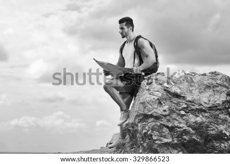 Black and white photo of a tourist with backpack standing on top of the mountain holding a map. Man with map exploring wilderness on trekking adventure. Tourist on the background of the sky