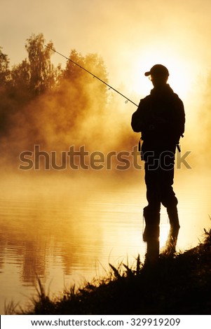 Fisher man fishing with spinning rod on a river bank at misty foggy sunrise