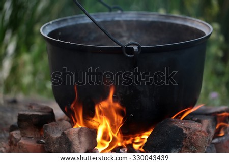 Closeup cooking in cauldron licked by flames on open fire fireplace made of bricks stones in field conditions over blurred nature background, horizontal picture