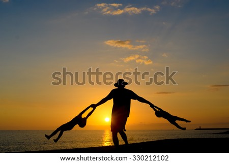 Picture of father holding and moving around two children in sunset scenic sunlights. Silhouette of happy family having fun on dramatic seascape background.