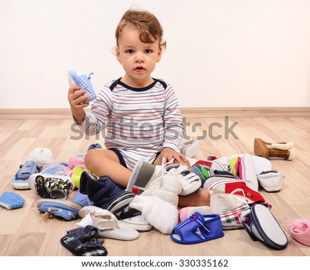 Toddler playing with a lot of baby shoes holding one shoe up. Untidy stack of child shoes thrown on the ground.  Royalty-Free Stock Photo #330335162