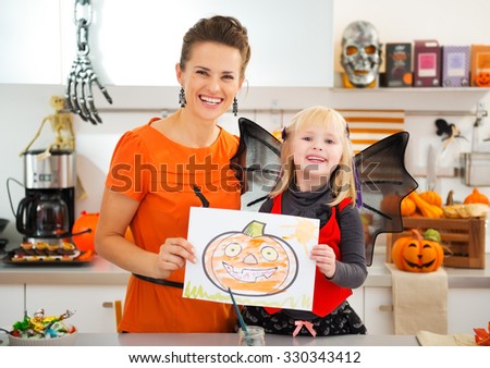 Happy halloween dressed blond girl with young mother showing self made painting of orange pumpkin Jack-O-Lantern in decorated kitchen. Traditional autumn holiday