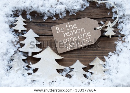 Brown Christmas Label With Ribbon On Wooden  Background With White Christmas Trees And Snow. Vintage Style. Label With English Quote Be The Reason Someone Smiles For Christmas Or Season Greetings