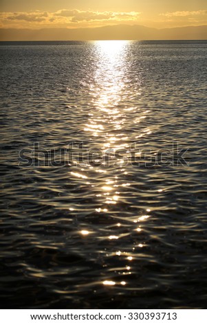 Marine sunset scene with lights reflecting at dark seaways in foreground and sun with halo glade shines over horizon in dusk against mountains silhouette with clouds background, vertical picture