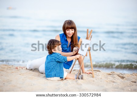 Woman and child in blue and white dress sit on the beach and draw on the easel