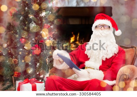 technology, holidays and people concept - man in costume of santa claus with tablet pc computer, gifts and christmas tree sitting in armchair over home fireplace and lights background