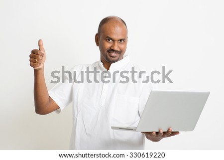 Portrait of mature casual business Indian man using laptop computer, looking at camera and thumb up, standing on plain background with shadow.