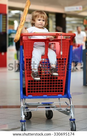 Closeup beautiful cute baby holding and biting French bread sitting in red and blue shopping trolley against supermarket full of people background, vertical picture