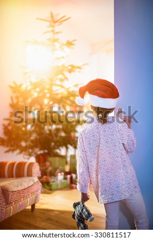 Christmas morning, a little girl in pajamas, wearing a Santa Claus hat opens the living room door and discovers the Christmas tree and many gift.The sun coming through the window gives cozy atmosphere