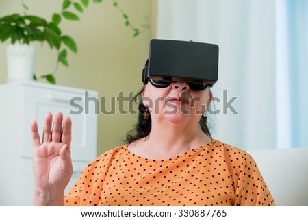Mature woman sitting on a couch at home with head-mounted display on. home interior on the background. She is touching the air with hand