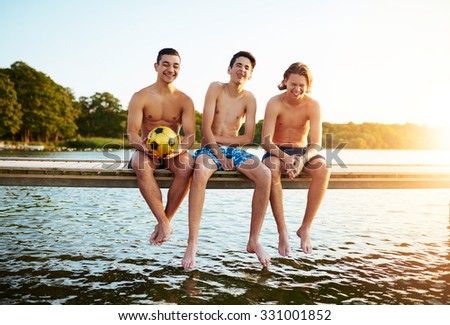 Three relaxed young multi ethnic teenage boys at a lake sitting side by side on a wooden jetty at sunset enjoying a summer camp together