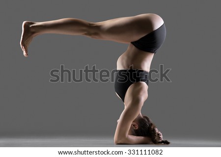 Beautiful young fit woman doing sport exercises, variation of supported headstand asana, salamba sirsasana posture with legs bent at right angle, full length, side view, studio shot on gray background