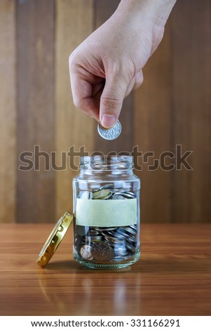 Image of hands putting coin into clear bottle with blank label against blurred wooden background. Saving concept.