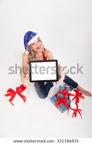 The beautiful blonde with big breasts is sitting on a white background and holds a a tablet in blue New Year's cap and smiling at the camera, selective focus on the tablet, picture with depth of field